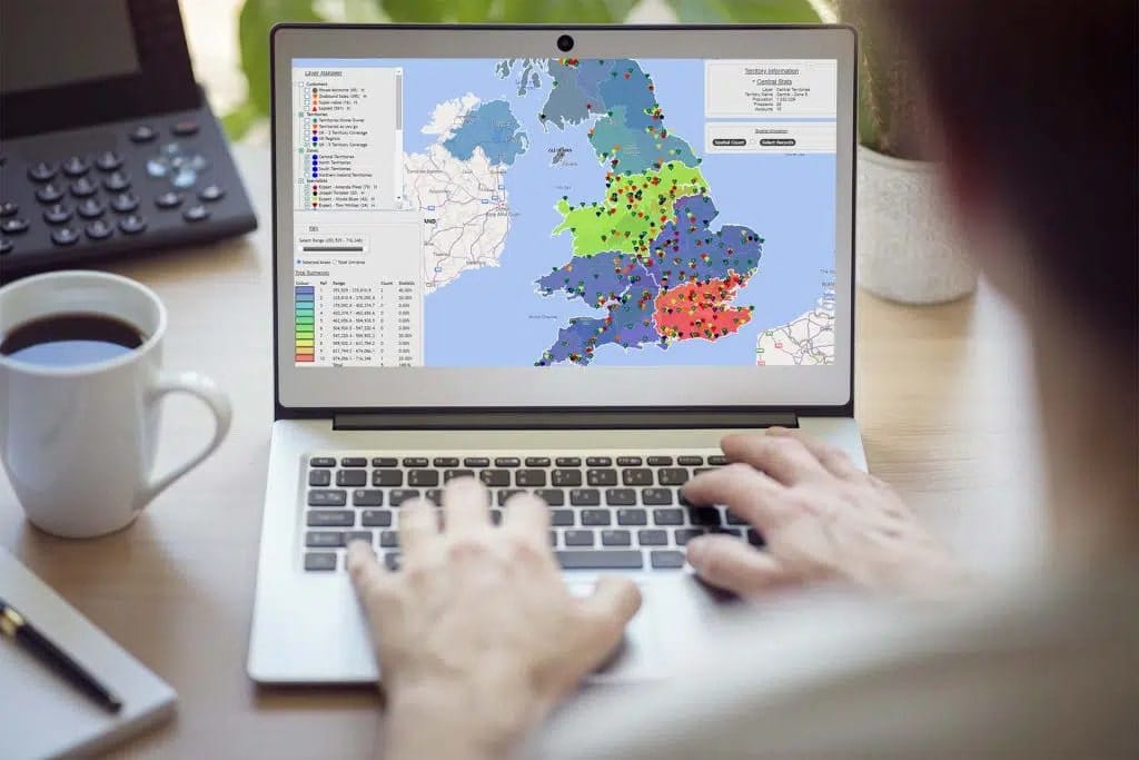 A person typing on a laptop whilst looking at a colour coded map of the UK to show how sales territory mapping can help you get an advantage on your competitors.